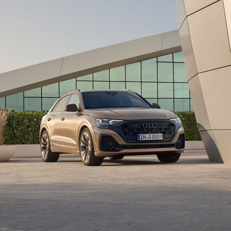 Der Q8 SUV schräg von vorn parkt vor einem modernen Gebäude. Eine bepflanzte Hecke trennt das Fahrzeug von dem dahinter liegenden, voll verglasten Gebäude.