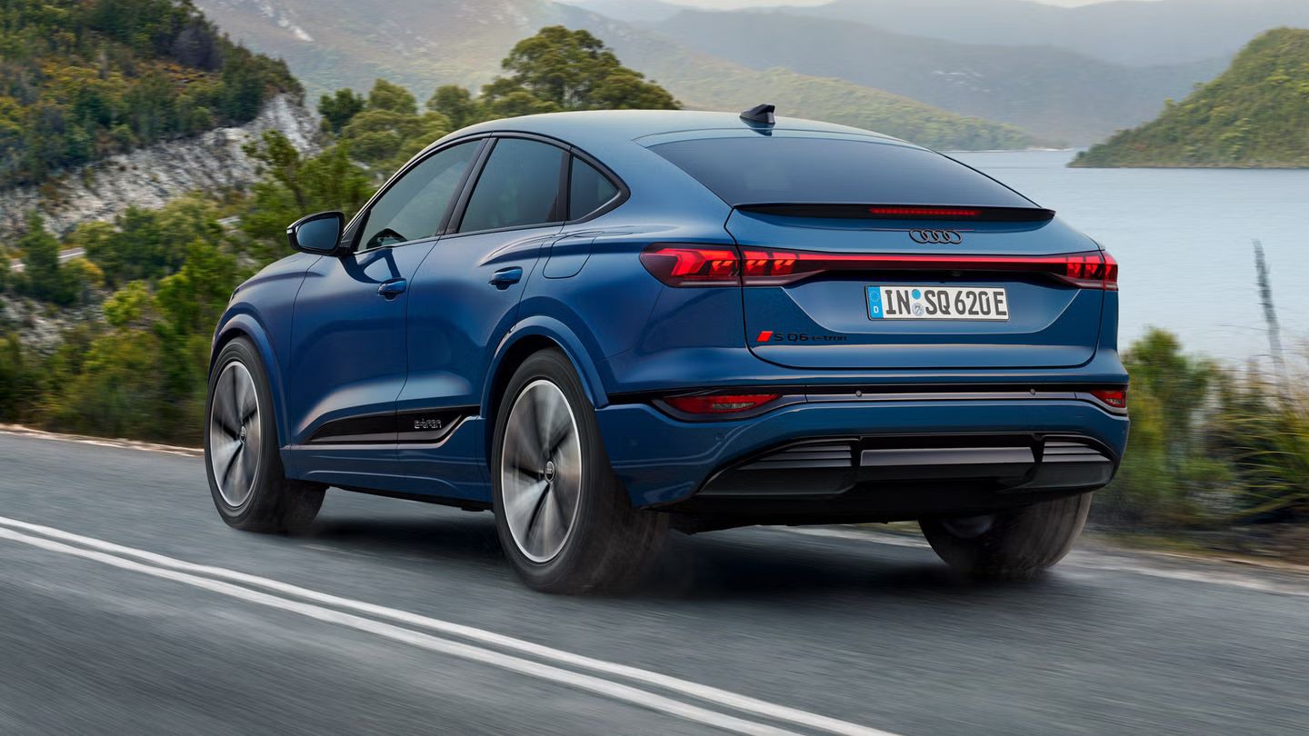 Schräge Heckaufnahme des SQ6 Sportback e-tron, welcher eine Straße an einem See in einer bergigen Landschaft entlangfährt.