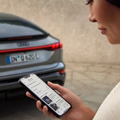 Frau schaut auf das Display ihres Mobiltelefons, im Hintergrund ist ein Audi zu sehen