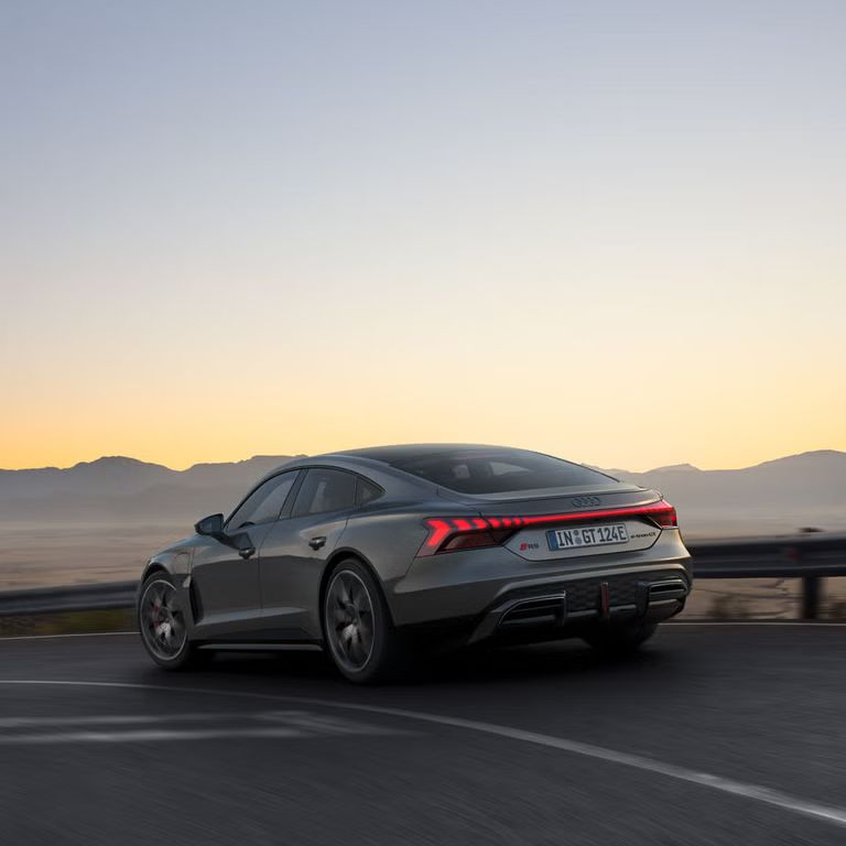 Ein grauer Audi RS e-tron GT fährt auf einer kurvigen Bergstraße bei Sonnenuntergang, mit leuchtenden Rücklichtern.