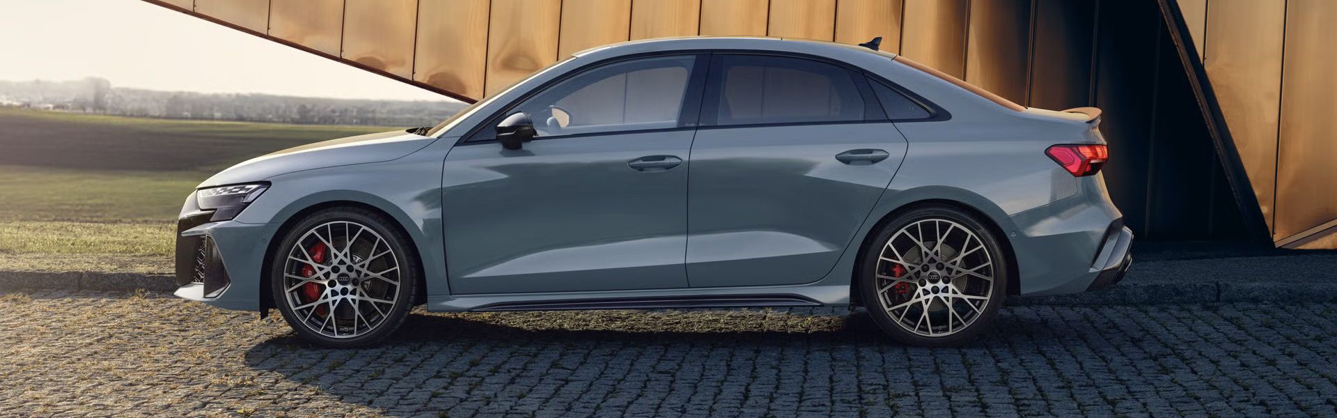 Die RS 3 Limousine von der Seite parkend auf Kopfsteinpflaster, im Hintergrund Umrisse eines modernen Gebäudes.