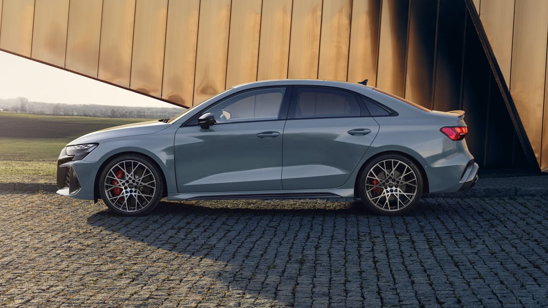 Die RS 3 Limousine von der Seite parkend auf Kopfsteinpflaster, im Hintergrund Umrisse eines modernen Gebäudes.