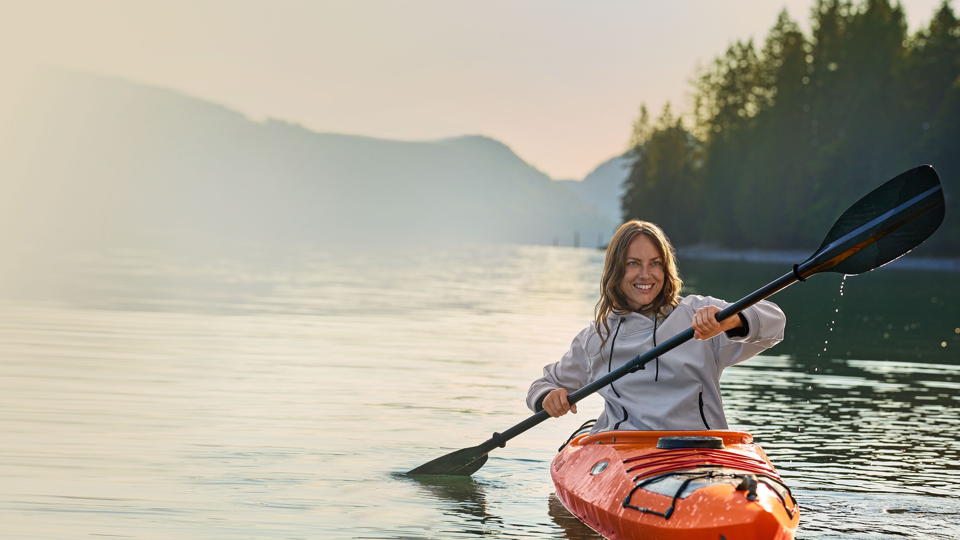 Eine Person in einem orangefarbenen Kajak paddelt auf einem ruhigen See, umgeben von bewaldeten Hügeln und Bergen in der Ferne.