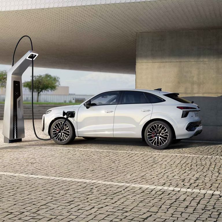 Auf dem Bild ist der Audi Q3 Sportback e-hybrid zu sehen. Dieser steht vor einem Gebäude mit einer Ladesäule und lädt.