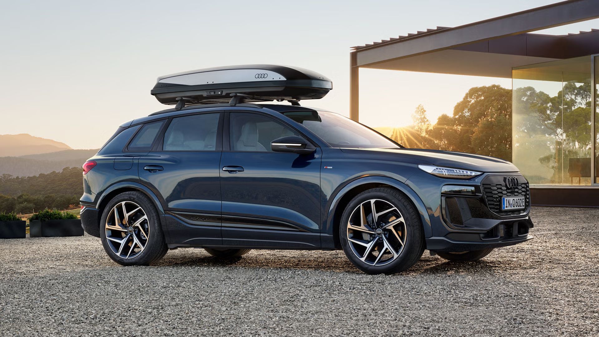 Der Q6 SUV e-tron mit Dachbox steht seitlich auf der Kiesfläche vor einem modernen Haus und wird dabei von der untergehenden Sonne angestrahlt.
