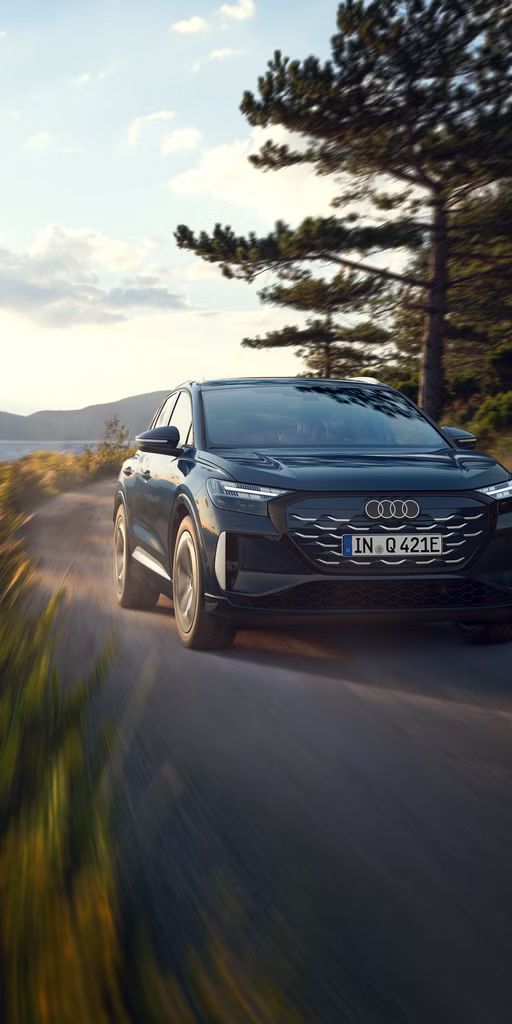 Der Q4 SUV e-tron schräg von vorn in voller Fahrt auf einer Landstraße an einem Waldsee, im Hintergrund eine bergige Landschaft.