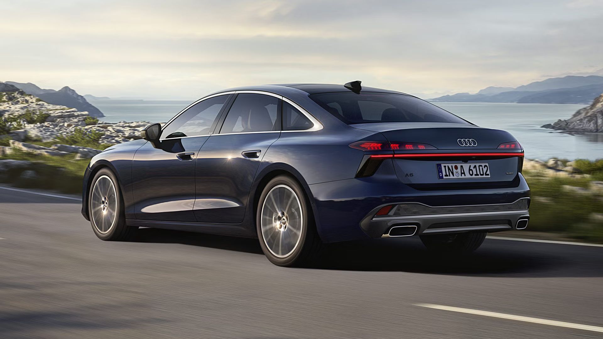 Die A6 Limousine in einer schrägen Heckperspektive, während sie auf einer kurvigen Straße entlang eines Fjords fährt. 