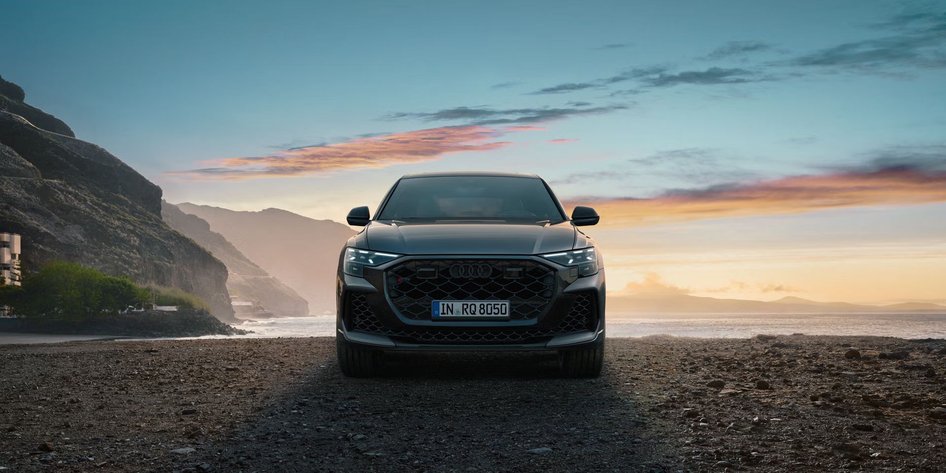 Der RS Q8 SUV von vorn an einem Strand auf Schotter, im Hintergrund das Meer und seitlich hervorragende Berge.