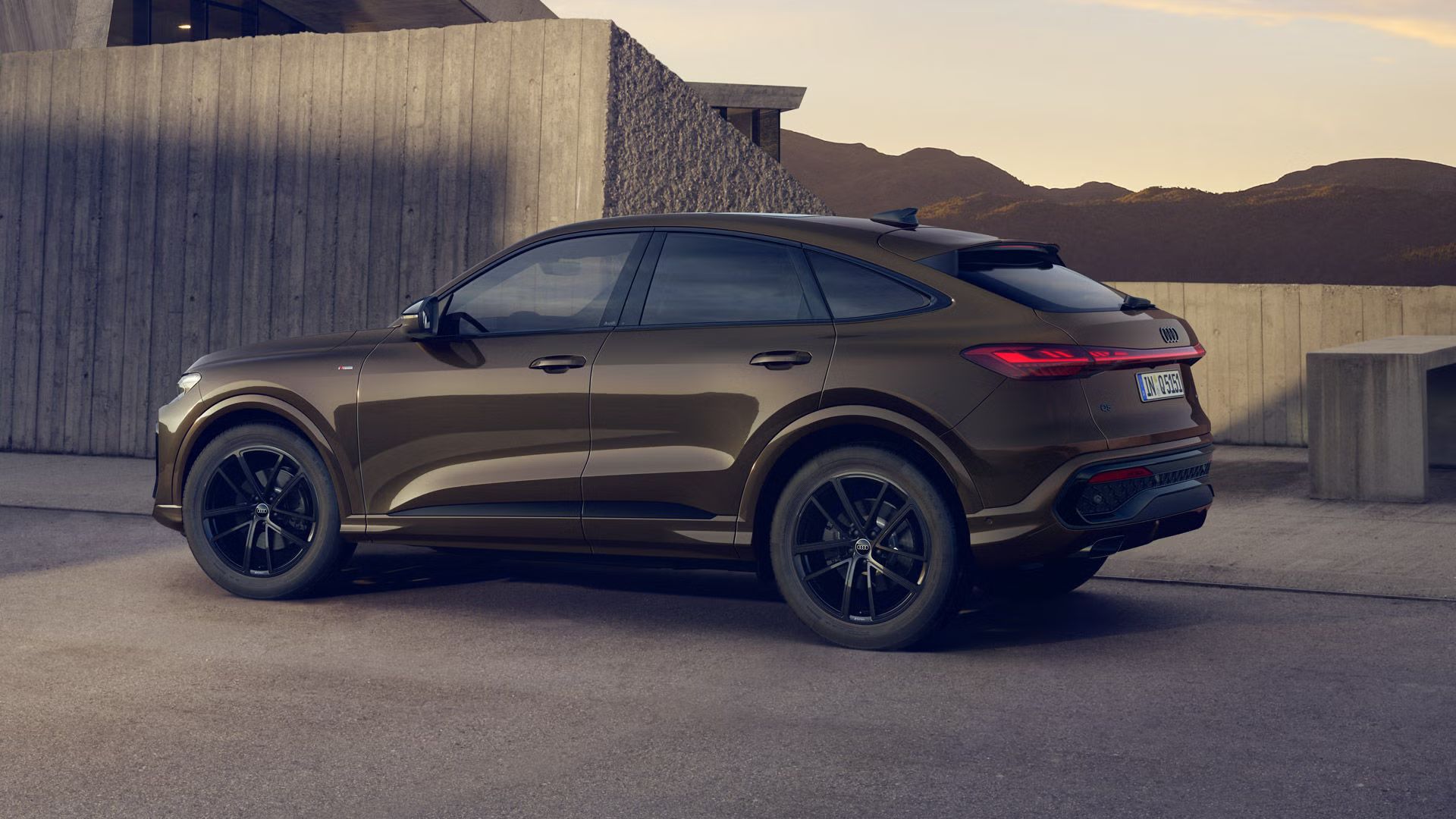 Der Q5 Sportback mit der Individuallackierung Teakbraun Metallic steht im Seitenprofil vor einem Gebäude. Im Hintergrund sind Berge zu erkennen.