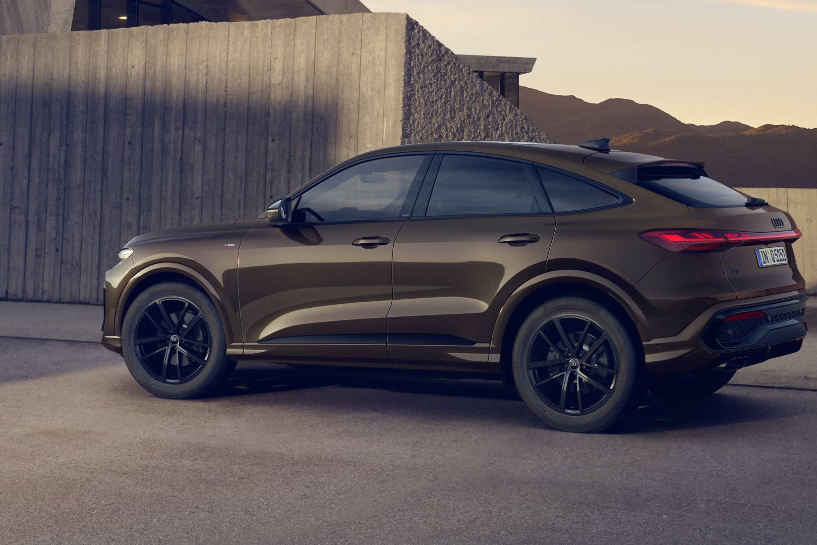Der Q5 Sportback mit der Individuallackierung Teakbraun Metallic steht im Seitenprofil vor einem Gebäude. Im Hintergrund sind Berge zu erkennen.