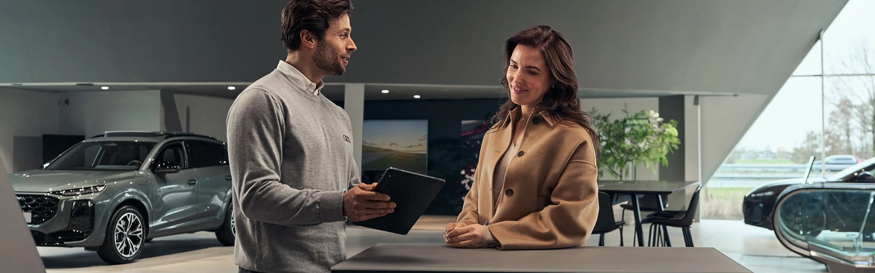 Ein Serviceberater im Gespräch mit einem Kunden am Informationsschalter, wobei er ein Tablet nutzt; im Hintergrund sind der Fahrzeugausstellungsbereich zu sehen.