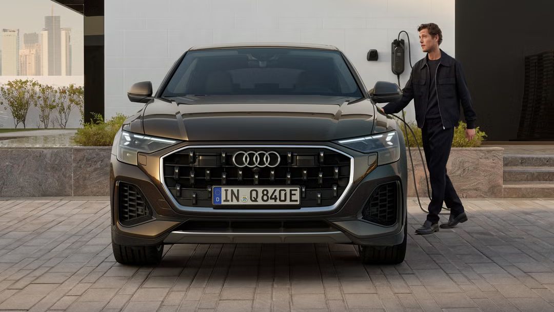 Frontansicht des Q8 SUV e-hybrid, dahinter ein modernes Gebäude. Neben den Fahrzeug steht ein modisch gekleideter junger Mann und hält den Stecker einer am Gebäude angebrachten Ladesäule in der Hand.