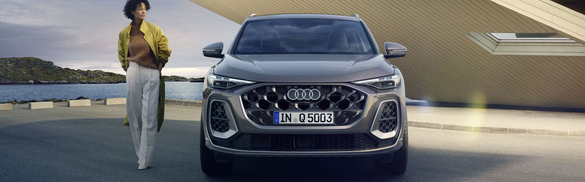 Der Q5 SUV im Frontprofil auf einem Parkplatz direkt an der Küste, dahinter ein kleines, futuristisches Haus. Neben dem Q5 SUV steht eine junge, modisch gekleidete Frau.	