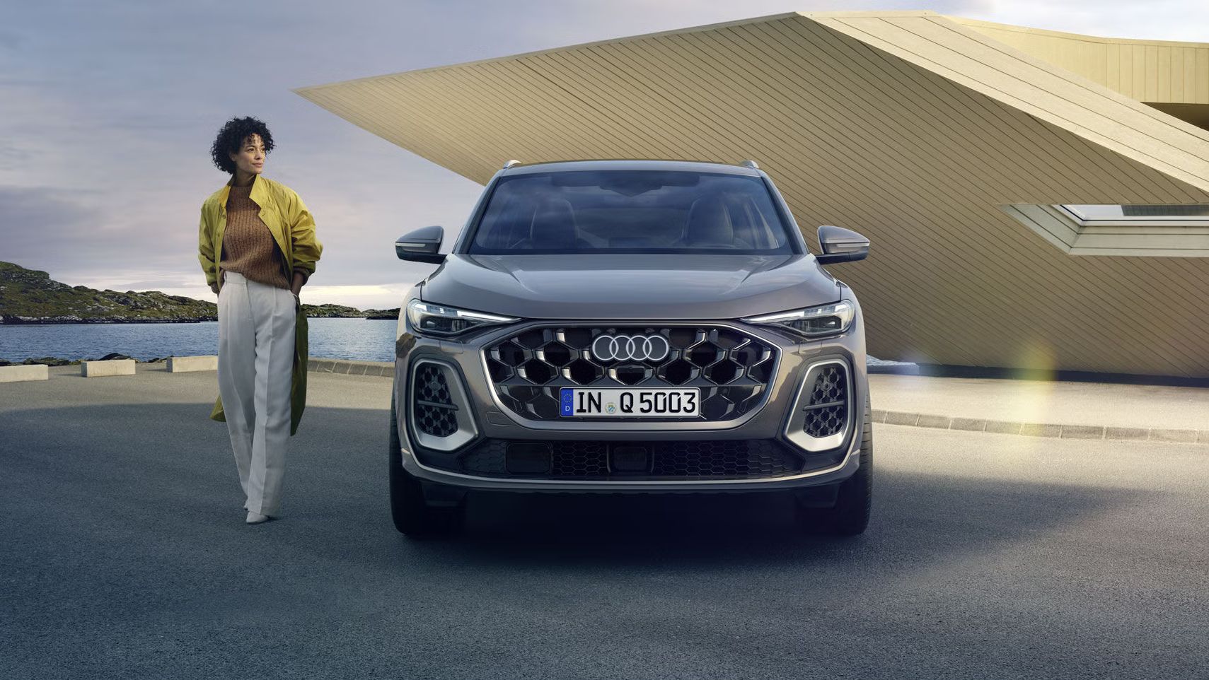 Der Q5 SUV im Frontprofil auf einem Parkplatz direkt an der Küste, dahinter ein kleines, futuristisches Haus. Neben dem Q5 SUV steht eine junge, modisch gekleidete Frau.	