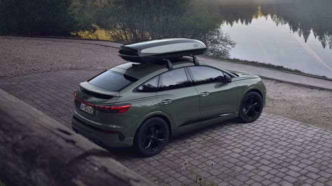 Der Q4 Sportback e-tron mit einer Dachbox schräg von hinten auf einem Parkplatz, an einem See parkend.