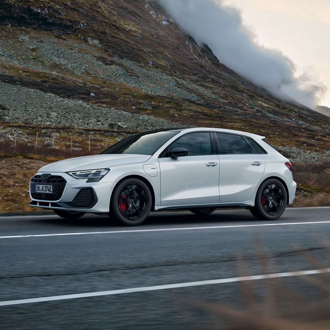Weißer Audi A3 Sportback e-hybrid fährt auf einer leeren Landstraße durch eine bergige Landschaft. Links erhebt sich ein steiler, felsiger Hang, aus dem sich Nebelschwaden lösen. Die Stimmung ist herbstlich und ruhig, mit gedämpften Farben und bewölktem Himmel.
