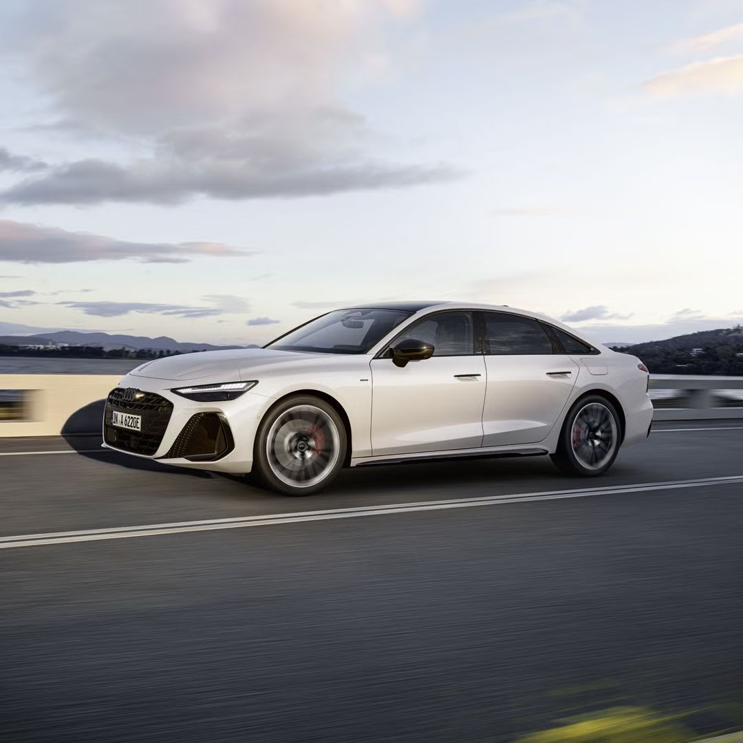 Seitliche Aufnahme der A6 Limousine e-hybrid in weiß, welche auf einer Straße entlang der Küste fährt. Am Steuer sitzt ein junger Mann.