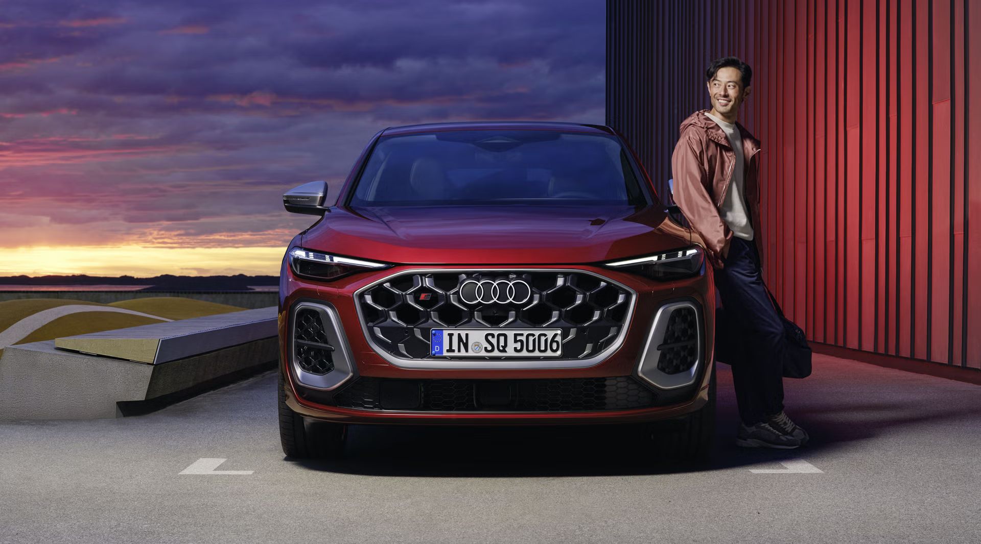 Der rote SQ5 Sportback steht auf einem Parkplatz parallel zu einem Hafen. Ein junger Mann lehnt sich an das Fahrzeug.