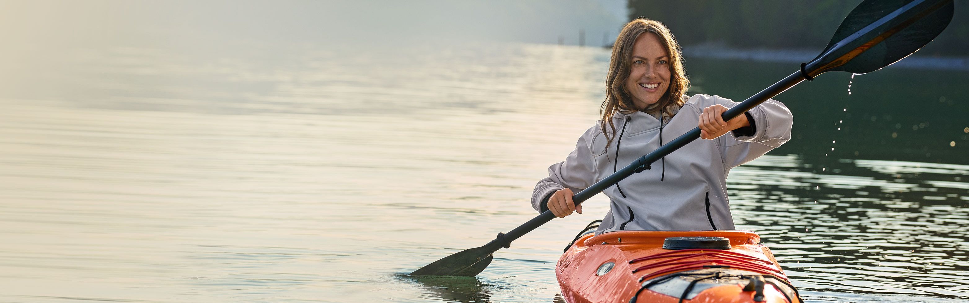 Eine Person in einem orangefarbenen Kajak paddelt auf einem ruhigen See, umgeben von bewaldeten Hügeln und Bergen in der Ferne.