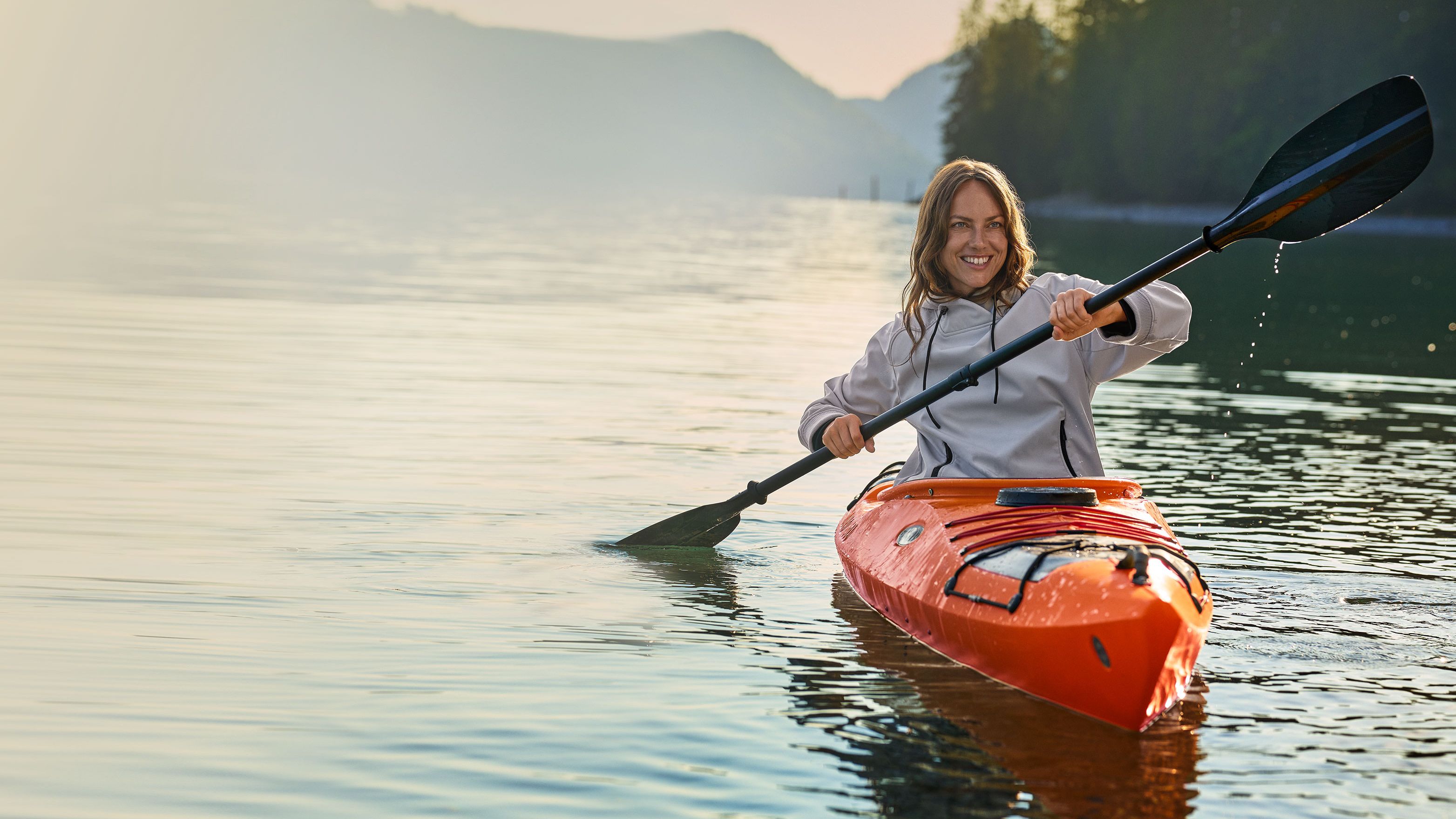 Eine Person in einem orangefarbenen Kajak paddelt auf einem ruhigen See, umgeben von bewaldeten Hügeln und Bergen in der Ferne.
