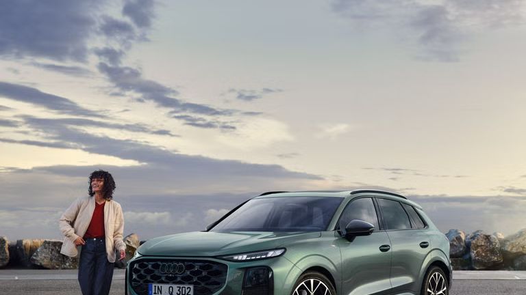 Seitliche, vordere Ansicht auf den geparkten Q3 SUV an einer Küstenstraße, daneben eine junge Frau. 