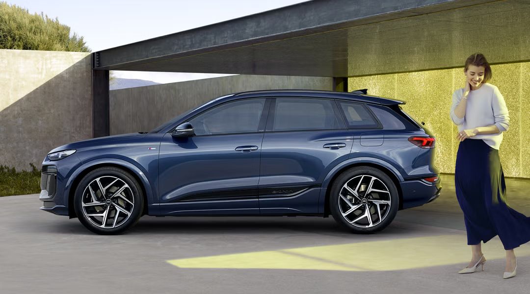 Der Q6 SUV e-tron in dunkelblau steht im Seitenprofil vor einem urbanen Haus. Im Vordergrund, leicht neben dem Auto steht eine junge, modische Frau.