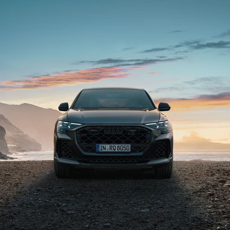 Der RS Q8 SUV von vorn an einem Strand auf Schotter, im Hintergrund das Meer und seitlich hervorragende Berge.