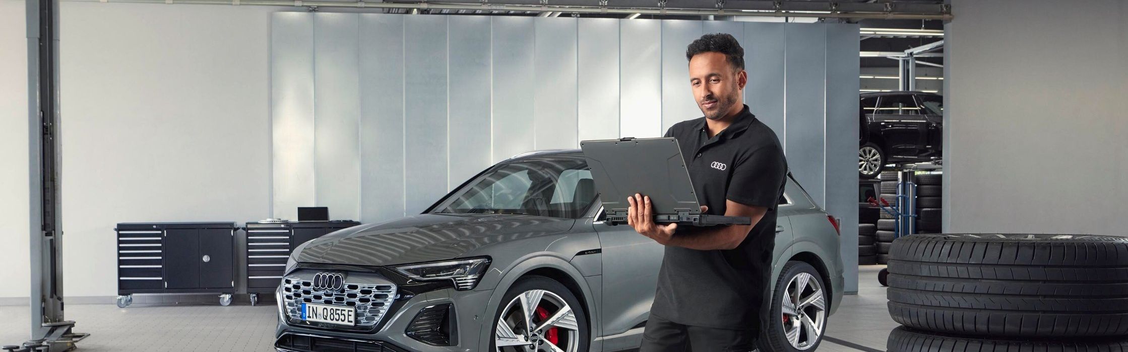 Ein Audi Service Mitarbeiter steht neben gestapelten Reifen und blickt auf sein Notebook.