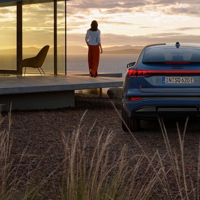 Heckprofil des SQ6 Sportback e-tron, auf Schotter parkend. Im Hintergrund das Meer bei Sonnenuntergang.