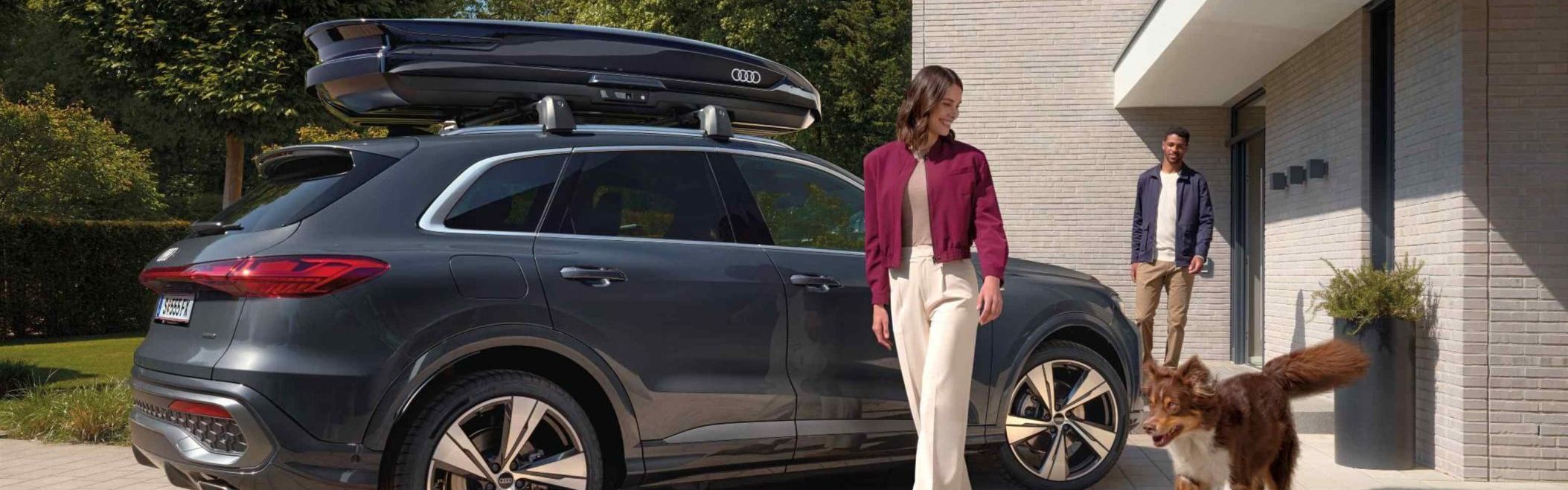Ein Australian Shepard läuft freudig neben einer lächelnden Frau und einem parkenden Audi Q5 mit Dachbox umher während ein Mann vor dem Hauseingang hinter den beiden steht