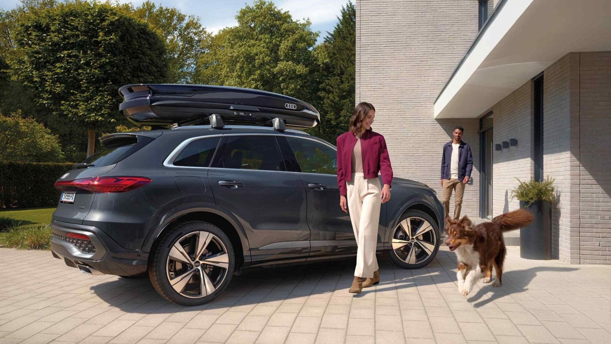 Ein Australian Shepard läuft freudig neben einer lächelnden Frau und einem parkenden Audi Q5 mit Dachbox umher während ein Mann vor dem Hauseingang hinter den beiden steht