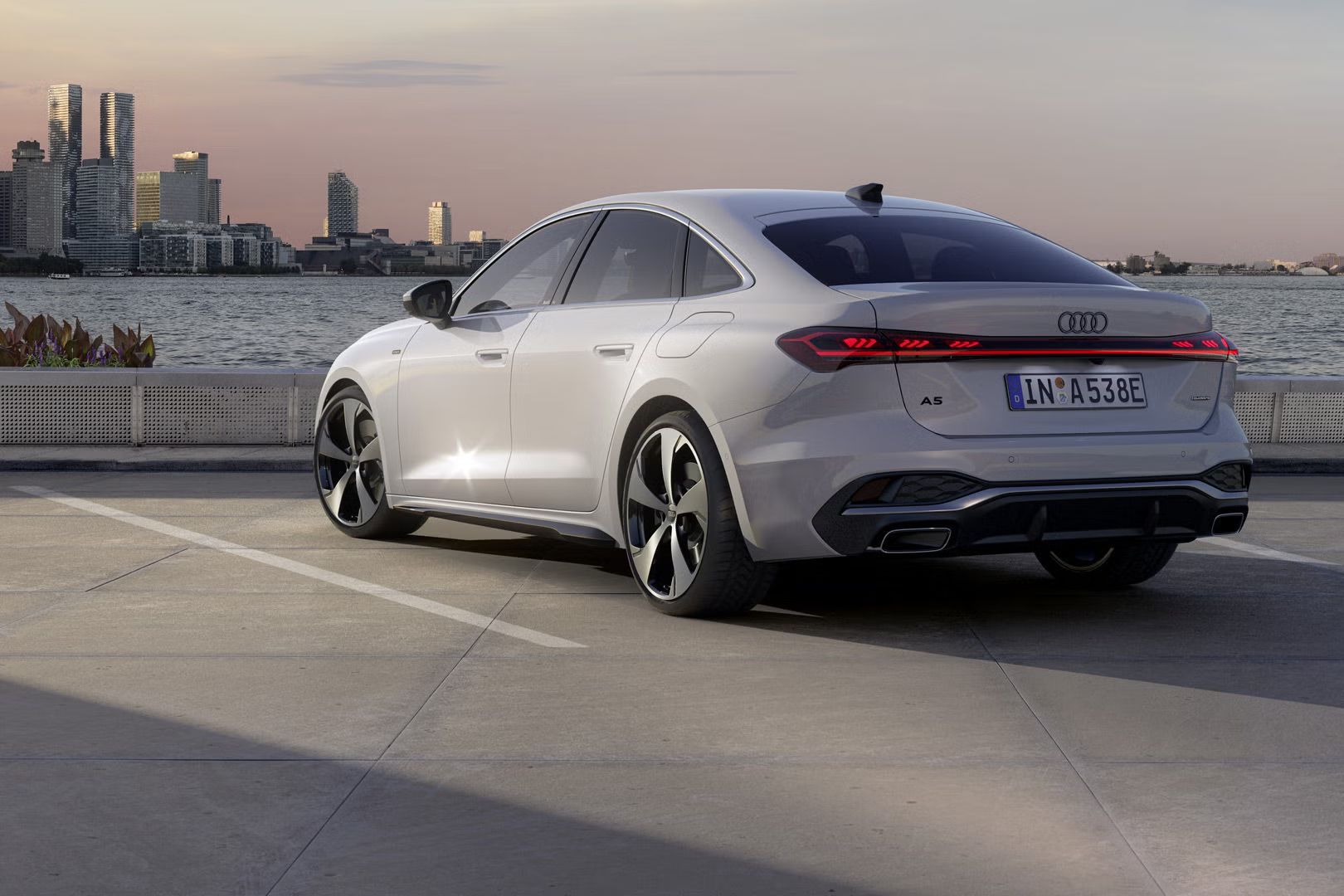 Die A5 Limousine e-hybrid steht im schrägen Heckprofil auf einem Parkplatz am Ufer eines Sees. Dahinter ist die Skyline einer Stadt bei Dämmerung zu sehen.