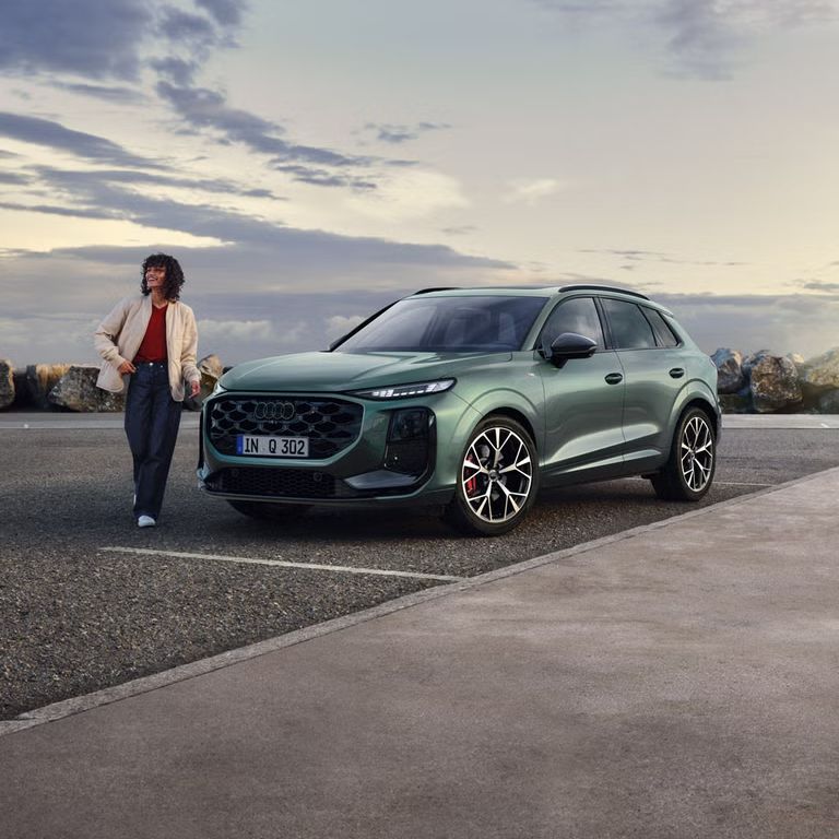 Seitliche, vordere Ansicht auf den geparkten Q3 SUV an einer Küstenstraße, daneben eine junge Frau. 