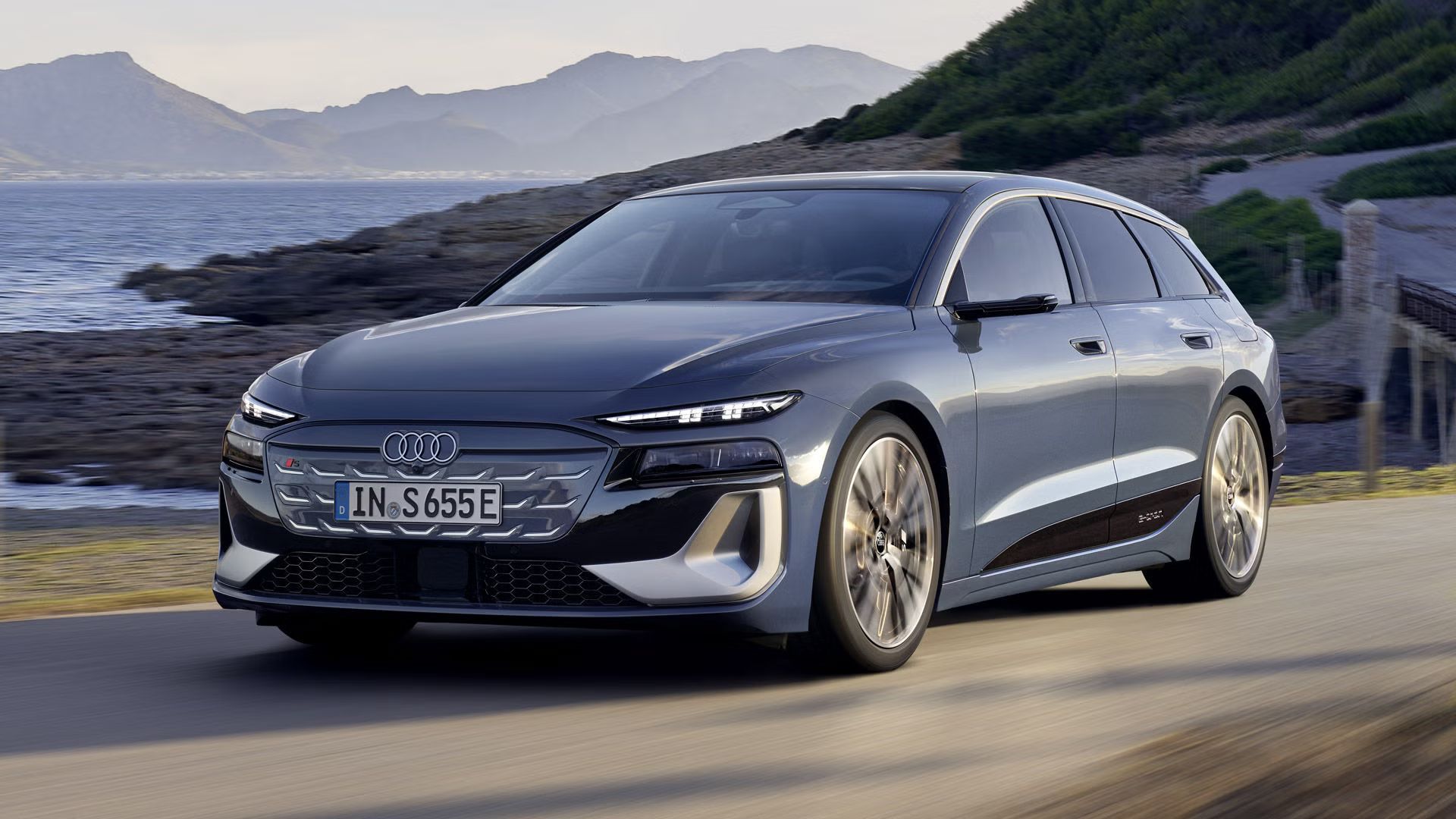 Der S6 Avant e-tron aus schräger Frontansicht, wie er eine Straße an der Küste entlangfährt. Im Hintergrund sind Berge zu sehen. 