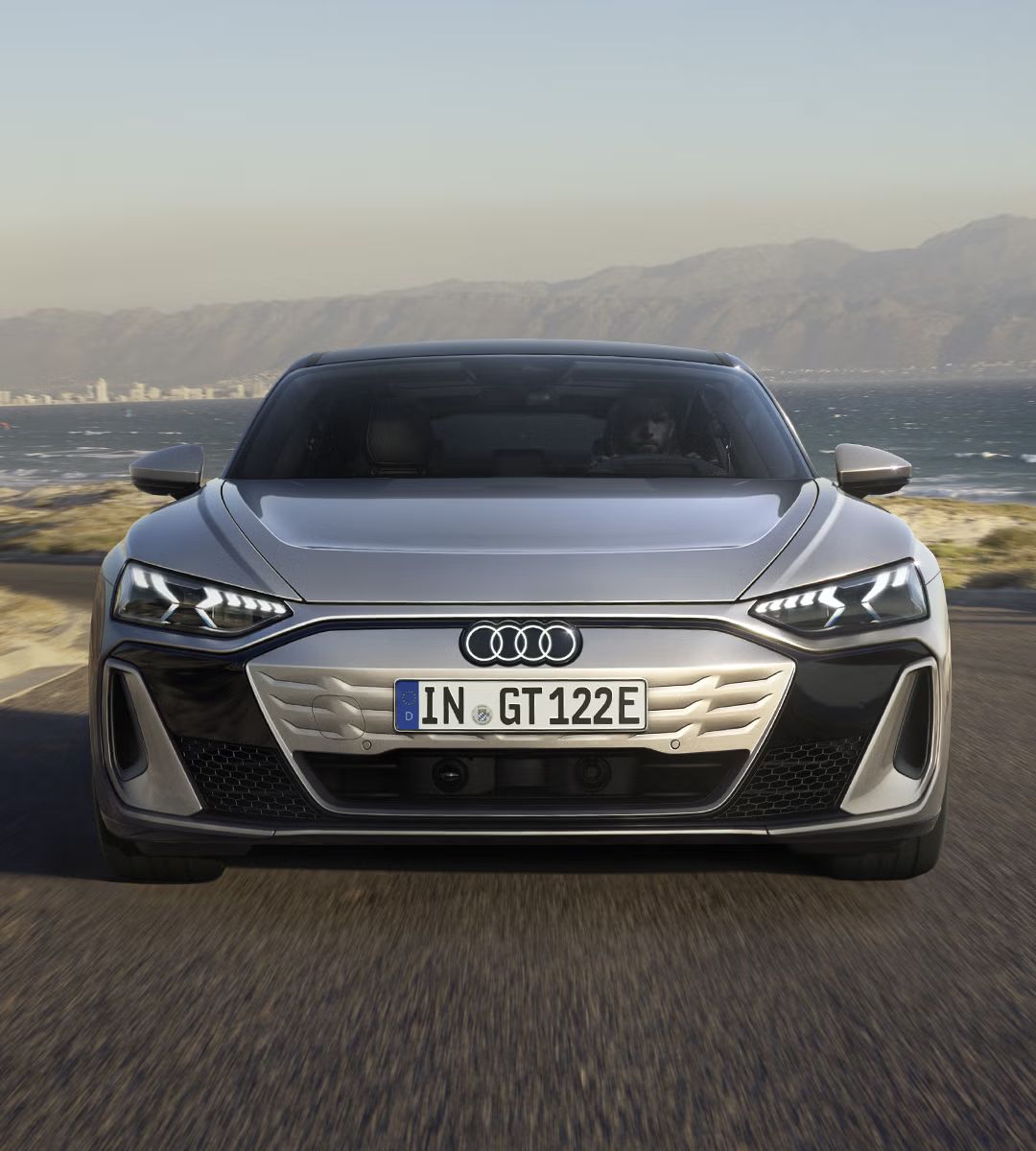Die Frontansicht des Audi S e-tron GT auf einer kurvigen Küstenstraße. Die markanten LED-Scheinwerfer und das geschlossene, aerodynamische Kühlergrill-Design sind erkennbar.