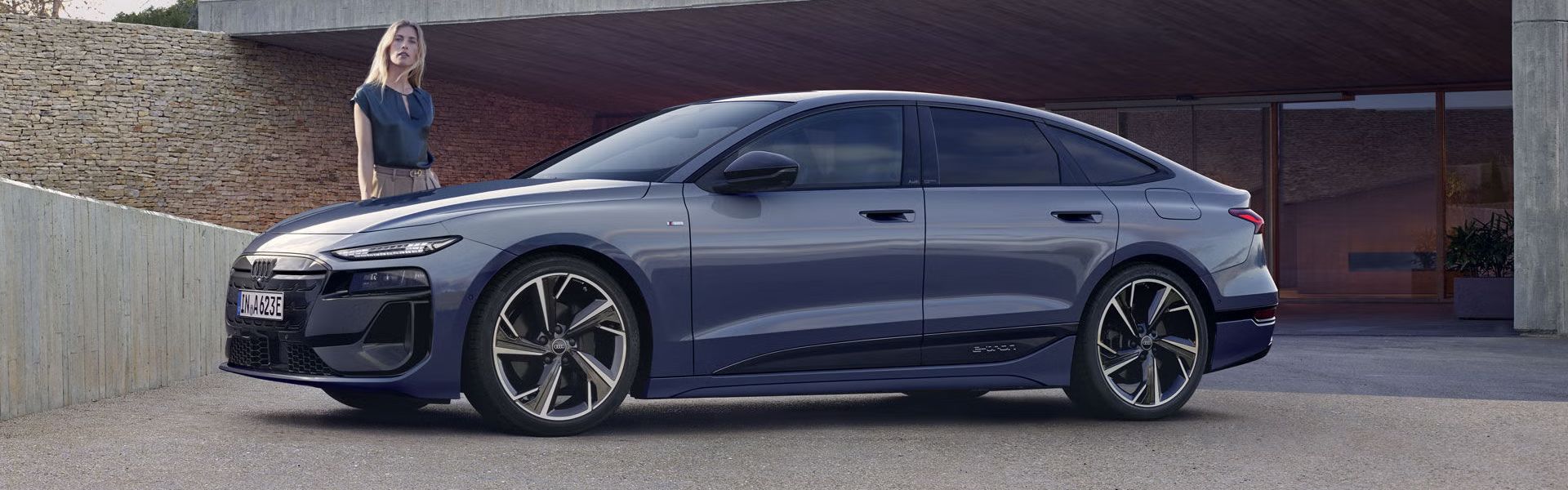 Der A6 Sportback e-tron steht im Seitenprofil vor dem Carport eines Hauses. Daneben steht eine junge Frau.