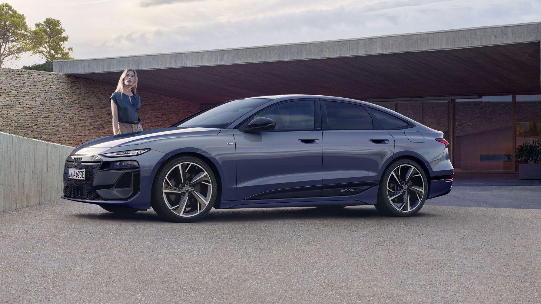 Der A6 Sportback e-tron steht im Seitenprofil vor dem Carport eines Hauses. Daneben steht eine junge Frau.