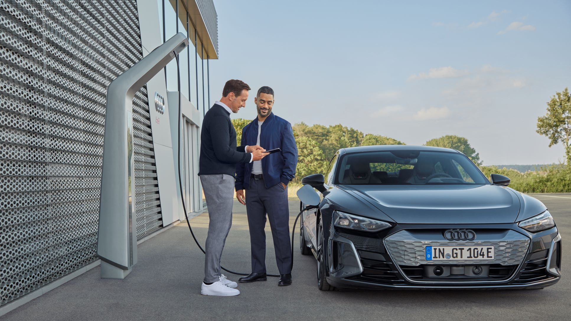 Zwei Männer unterhalten sich über eine Information am Smartphone, während ein Audi an einer Ladesäule aufgeladen wird.