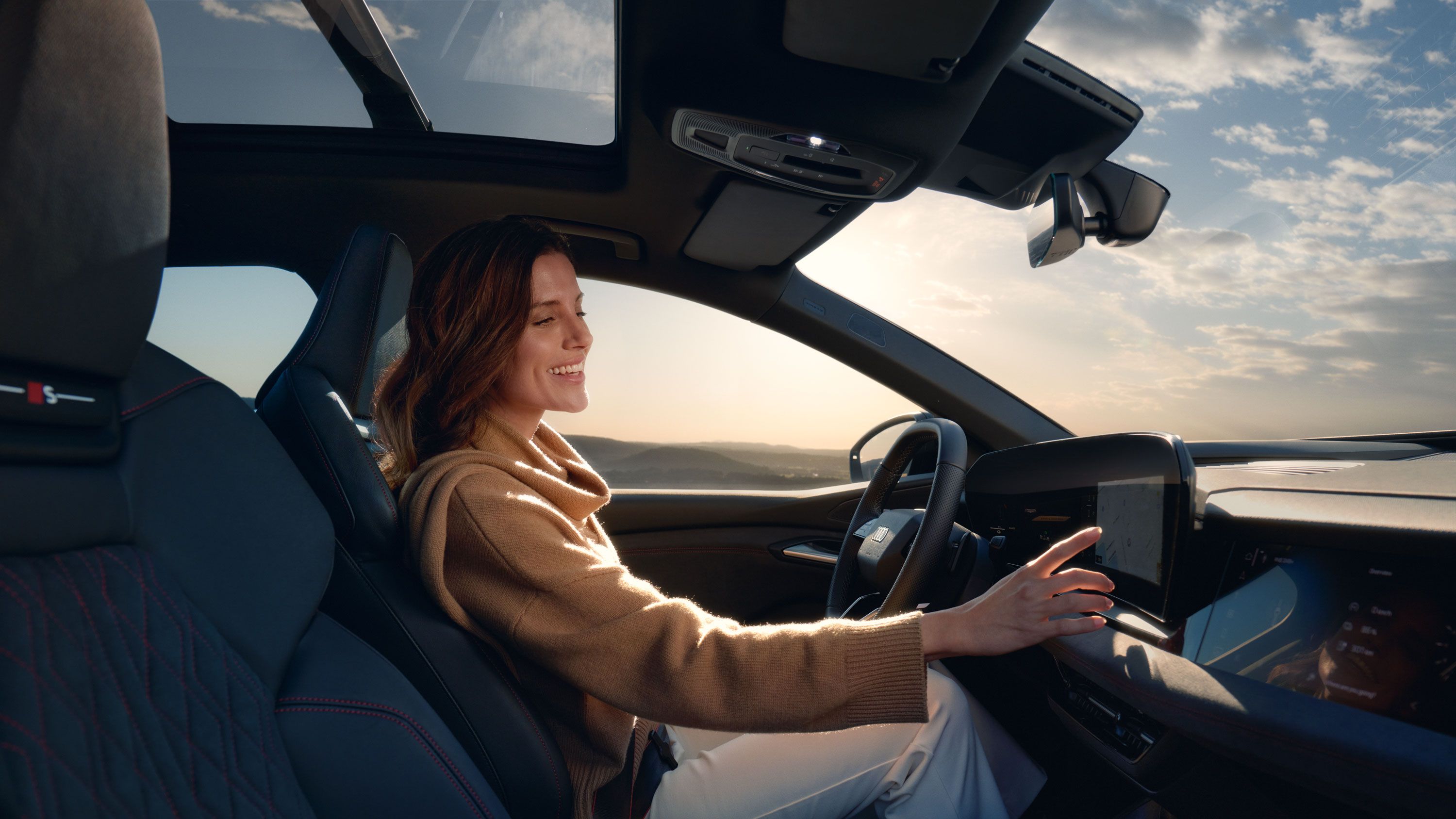 Junge Frau sitzt lächelnd am Steuer eines Audi und bedient den Touchscreen des MMI. Im Hintergrund ist eine weite Landschaft unter leicht bewölktem Himmel zu sehen.