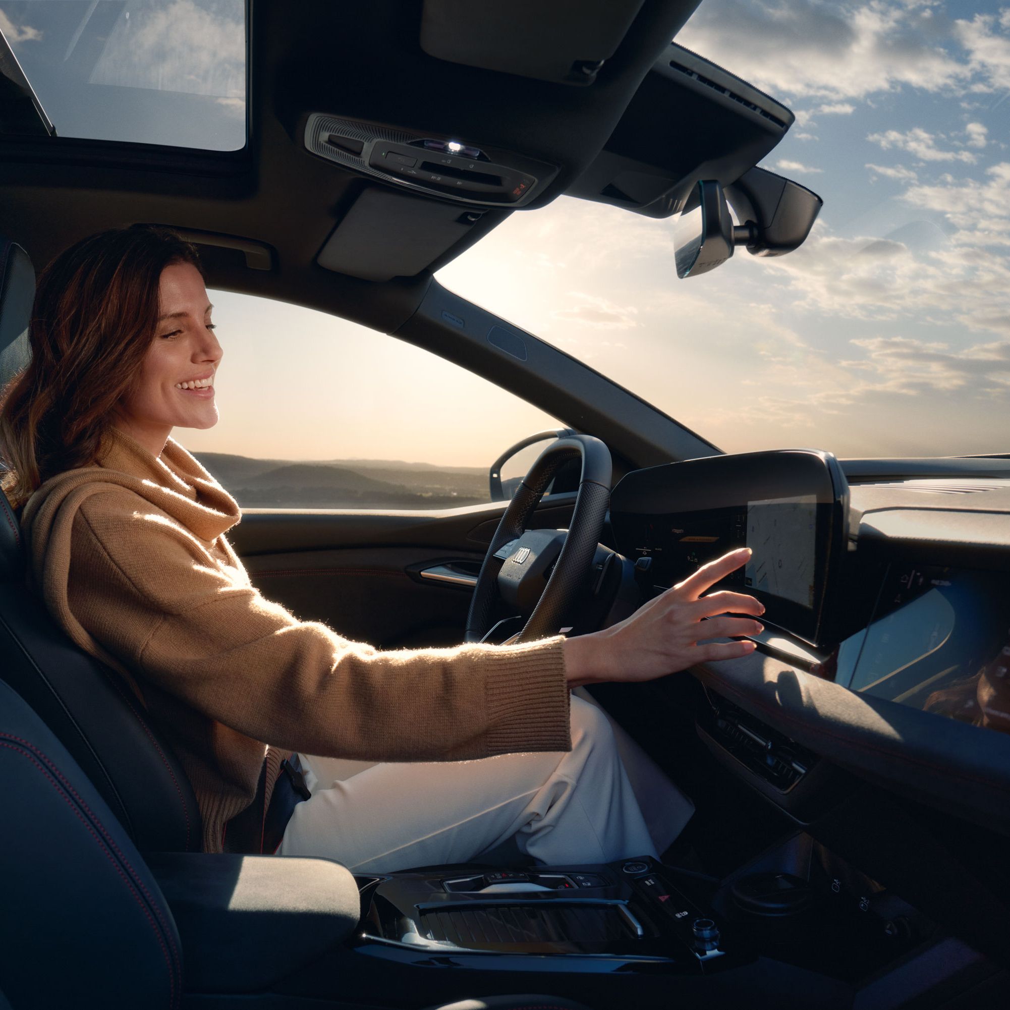Junge Frau sitzt lächelnd am Steuer eines Audi und bedient den Touchscreen des MMI. Im Hintergrund ist eine weite Landschaft unter leicht bewölktem Himmel zu sehen.