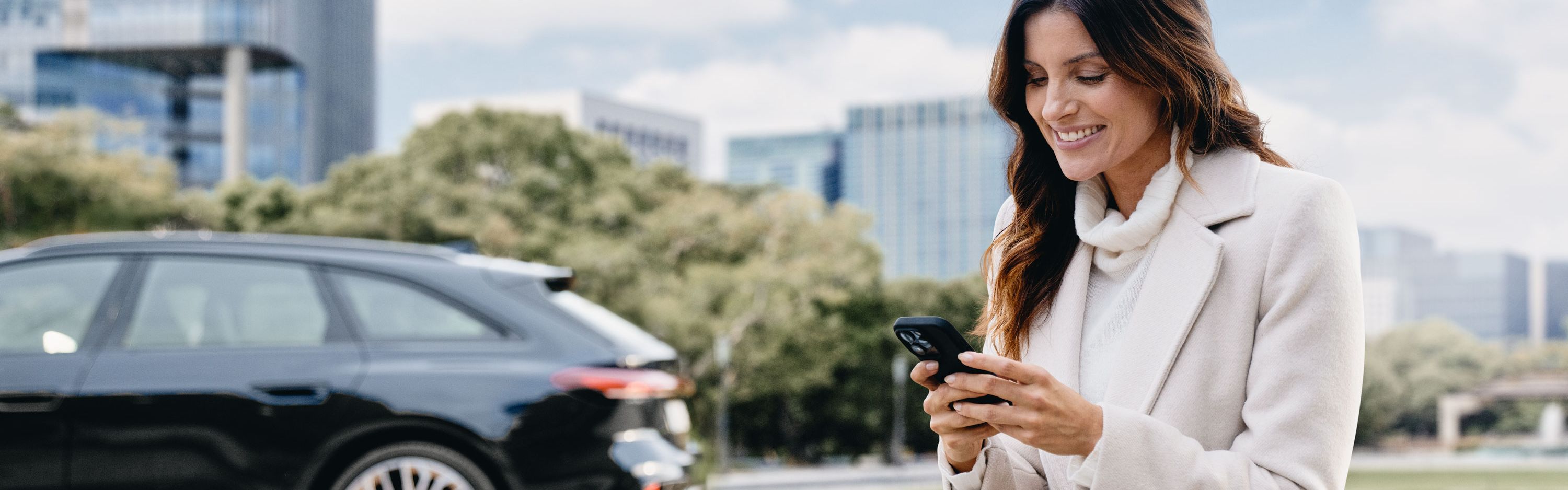 Eine Frau sitzt im Freien auf einer Steinmauer und schaut lächelnd auf ihr Smartphone. Im Hintergrund ist ein schwarzer Audi vor modernen Bürogebäuden und Bäumen geparkt.