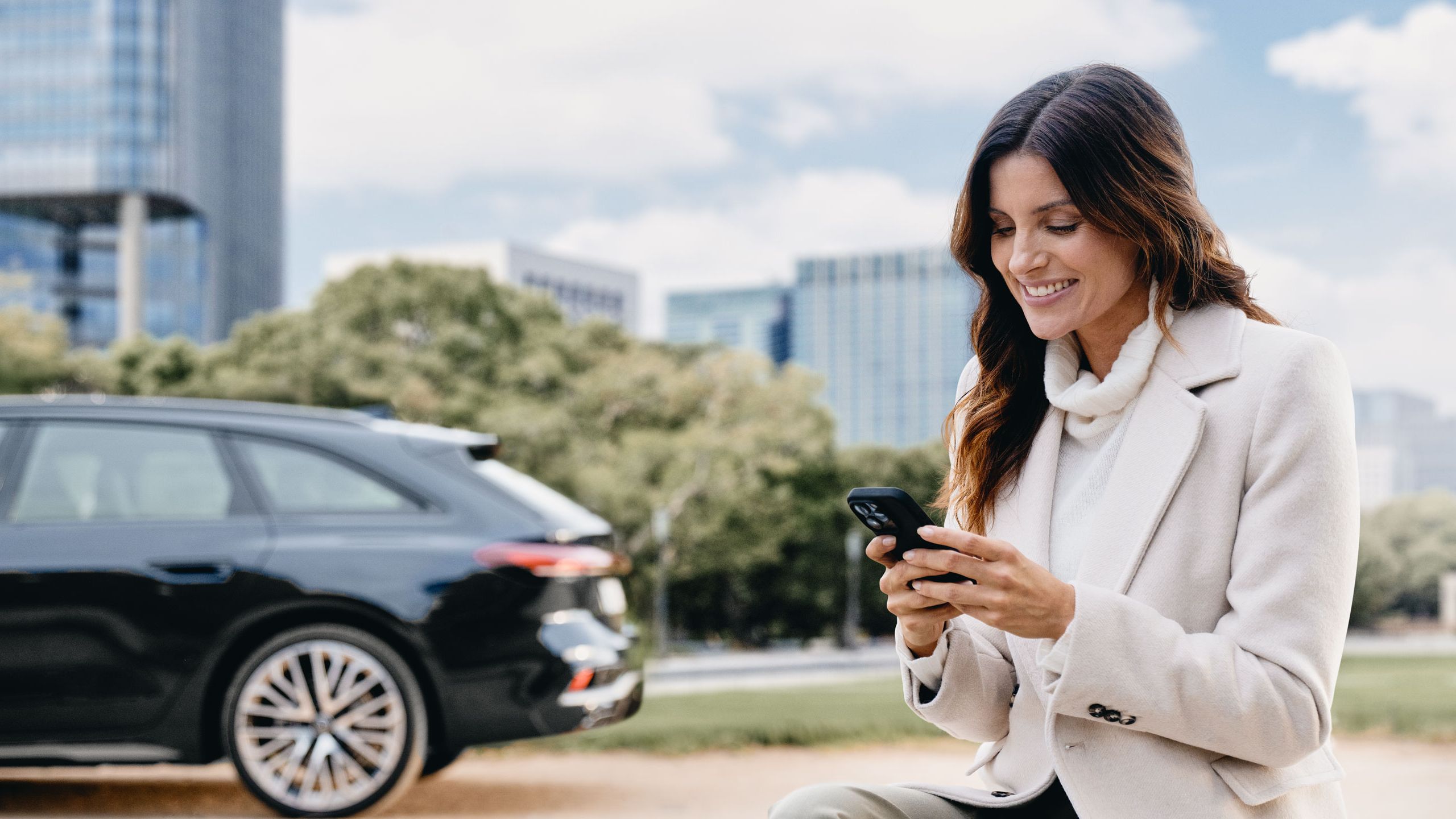 Eine Frau sitzt im Freien auf einer Steinmauer und schaut lächelnd auf ihr Smartphone. Im Hintergrund ist ein schwarzer Audi vor modernen Bürogebäuden und Bäumen geparkt.