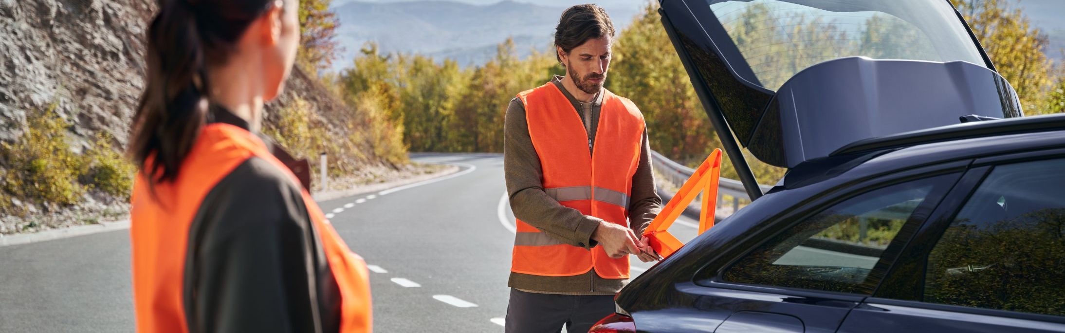 Zwei Personen in orange Warnwesten stehen neben ihrem Audi auf einer kurvenreichen Straße. Eine Person holt ein Warndreieck aus dem Kofferraum, während die andere Person in deren Richtung blickt.