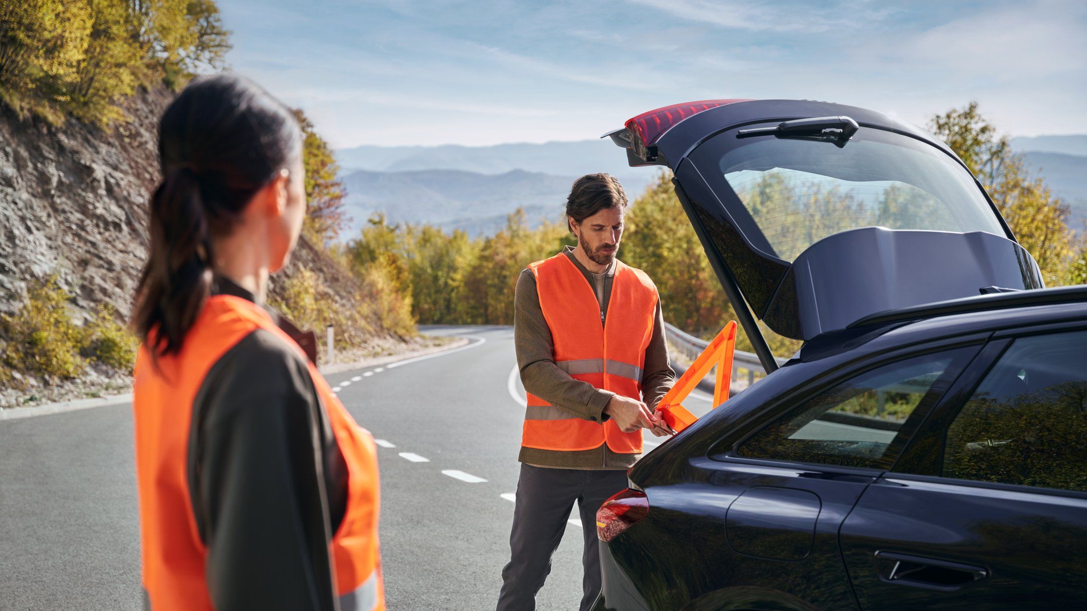 Zwei Personen in orange Warnwesten stehen neben ihrem Audi auf einer kurvenreichen Straße. Eine Person holt ein Warndreieck aus dem Kofferraum, während die andere Person in deren Richtung blickt.