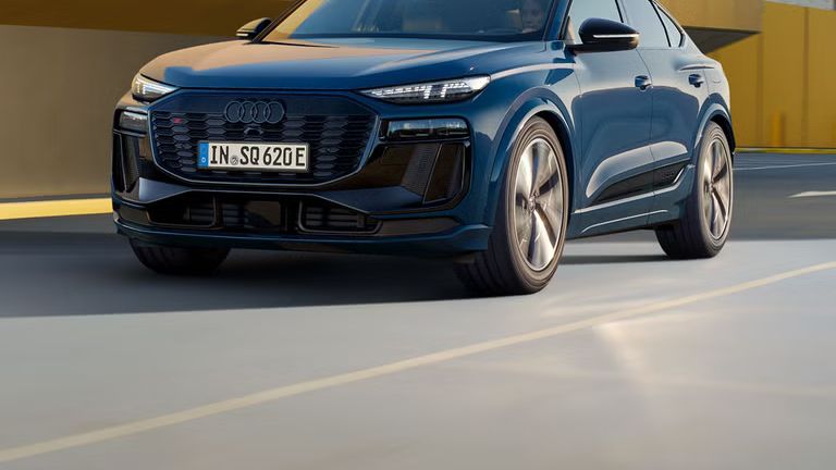 Schräges Frontprofil des SQ6 Sportback e-tron, welcher auf einer Straße durch die Stadt fährt. Im Hintergrund sieht man eine gelbe Gebäudefassade.