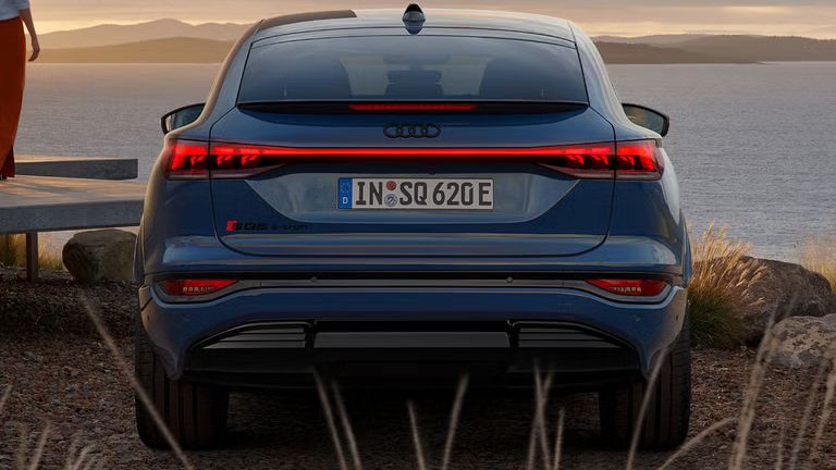 Heckprofil des SQ6 Sportback e-tron, welcher vor einem modernen Haus direkt an der Küste steht. Auf der Veranda steht eine junge Frau und schaut in Richtung des Wassers.