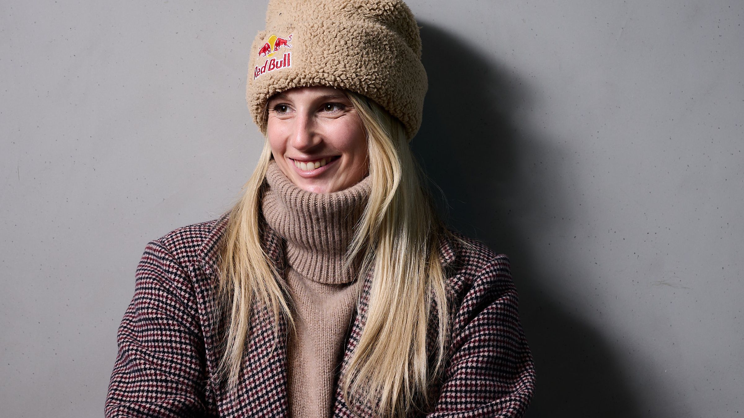 Anna Gasser trägt eine beige Mütze und einen karierten Mantel und lehnt an einer grauen Wand. 