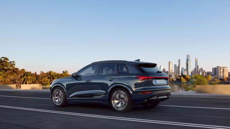 Seitenprofil des Q6 SUV e-tron, welcher auf einer Straße an einem Fluss fährt. Im Hintergrund ist die Skyline einer Großstadt zu sehen.