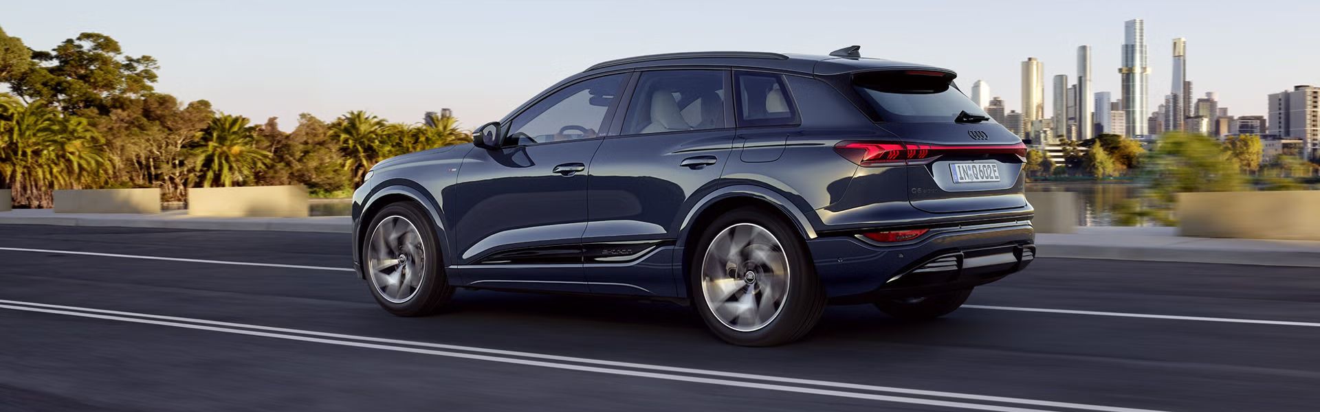 Seitenprofil des Q6 SUV e-tron, welcher auf einer Straße an einem Fluss fährt. Im Hintergrund ist die Skyline einer Stadt zu sehen.