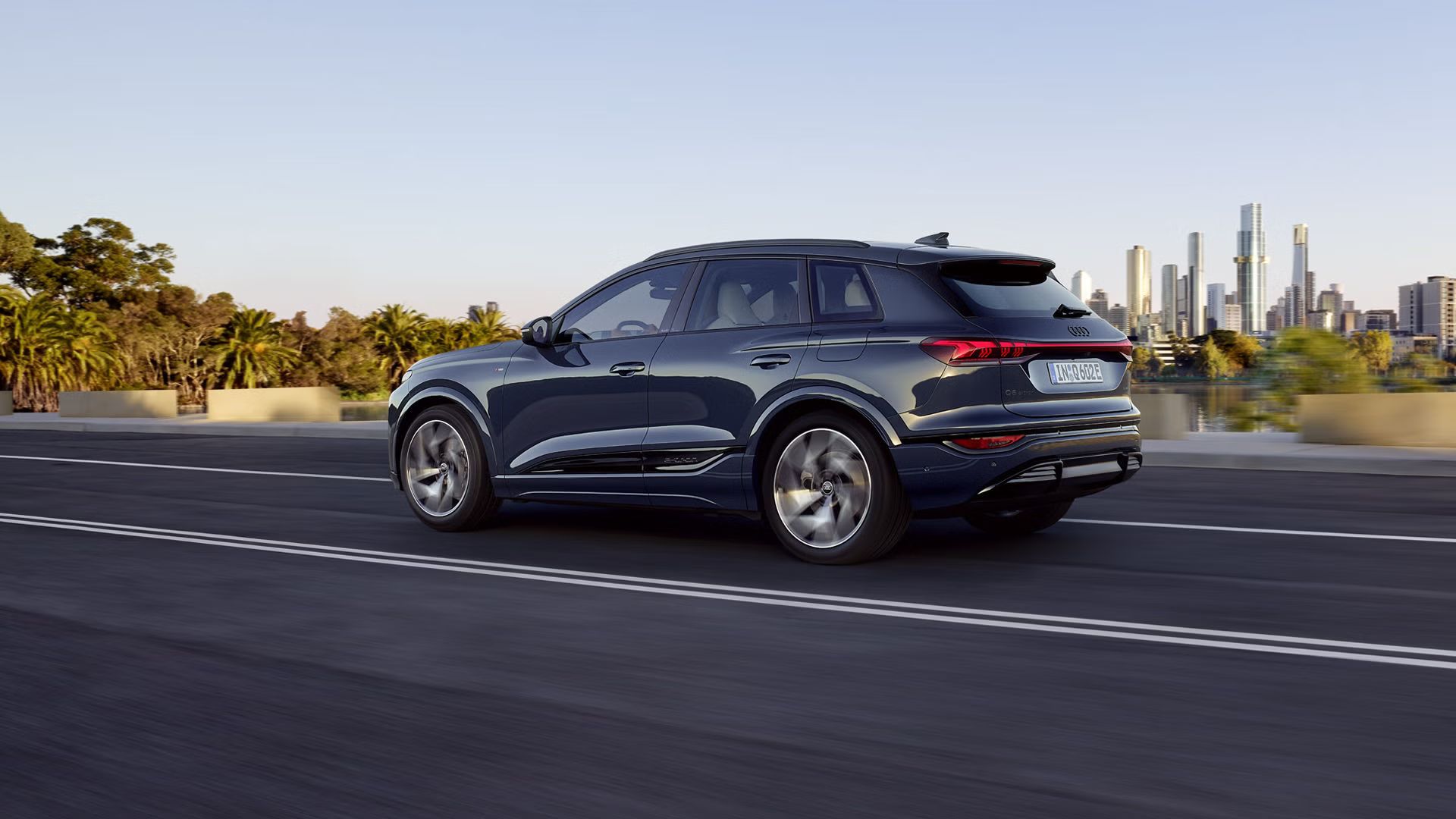 Seitenprofil des Q6 SUV e-tron, welcher auf einer Straße an einem Fluss fährt. Im Hintergrund ist die Skyline einer Stadt zu sehen.